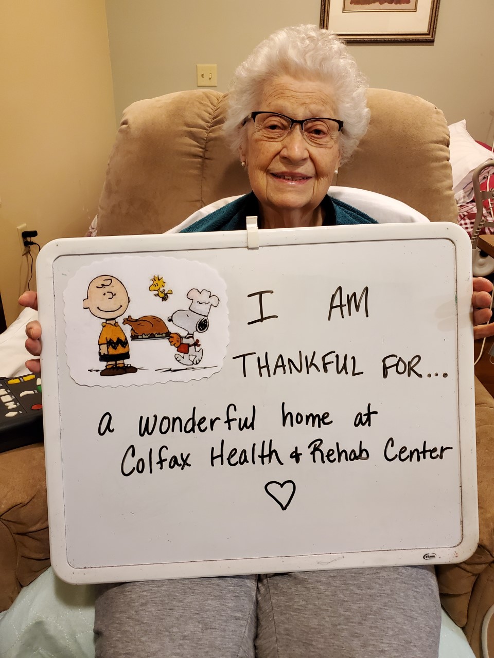 A person holding a whiteboard with "I AM THANKFUL FOR..." message, mentioning Colfax Health & Rehab Center, seated comfortably indoors with cheerful decor.