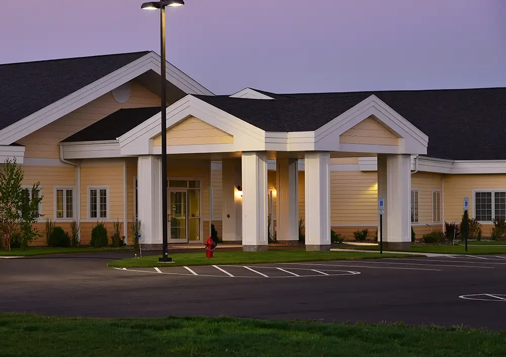 Modern beige building with white columns, a spacious parking lot, and surrounding greenery, under twilight sky. No people visible.