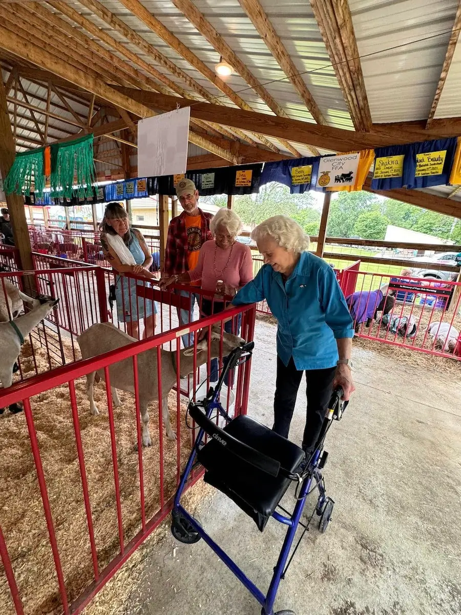 Four people interacting with goats in a barn. One person uses a walker. Colorful banners hang from the ceiling.