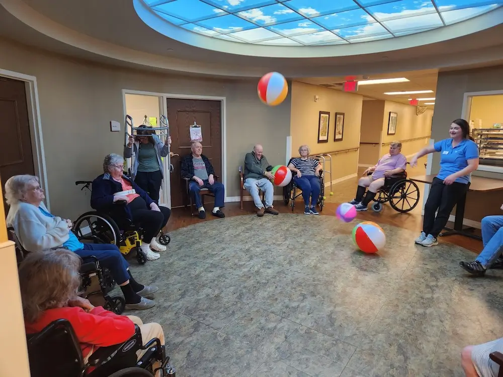 A group of people, some in wheelchairs, enjoy a beach ball activity in a room with a bright, sky-like ceiling.