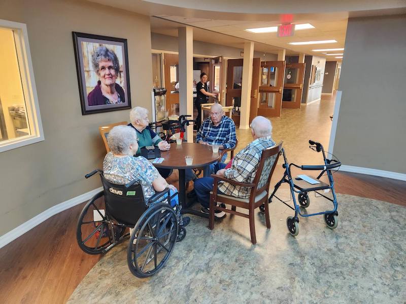 Four people sit around a table in a care facility, with mobility aids nearby and a large portrait on the wall.