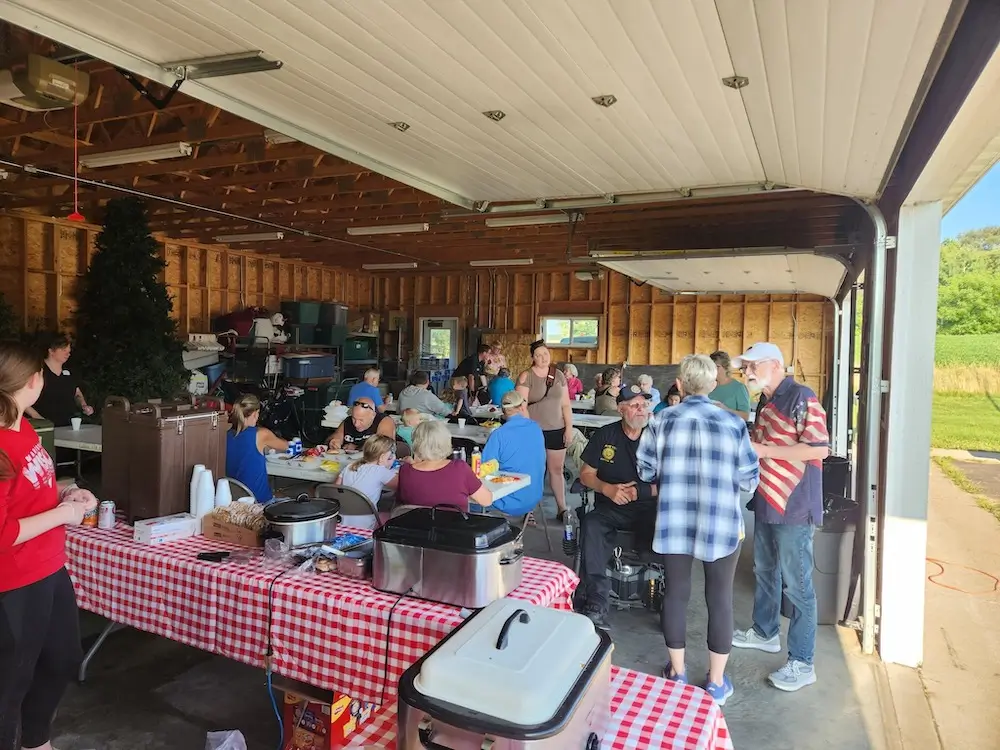 A group of people socializing in a garage, dining at tables with red checkered tablecloths, surrounded by various storage items.