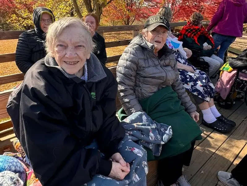 A group of people sit on a wooden bench outdoors, wearing jackets. Autumn leaves surround them, creating a colorful backdrop.