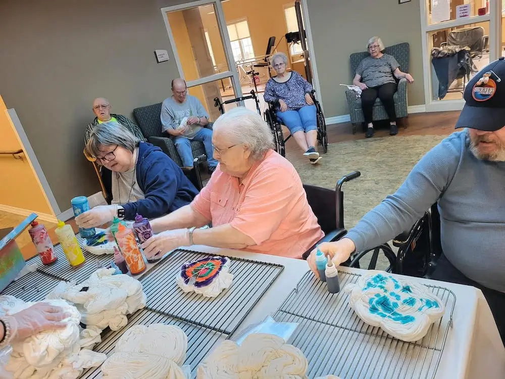 Several people in a room are sitting at a table, engaging in a tie-dye activity with colorful bottles and white fabric.
