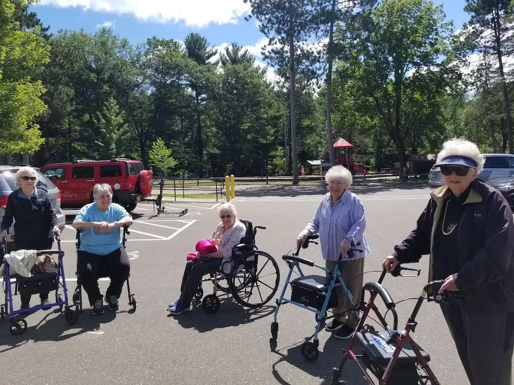 Five people with walkers are gathered in a sunlit parking lot by a park, with trees and playground equipment in the background.
