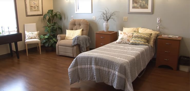 Cozy bedroom with a single bed, decorative pillows, chair, plants, and small table. Walls feature framed art; soft lighting sets a warm ambiance.