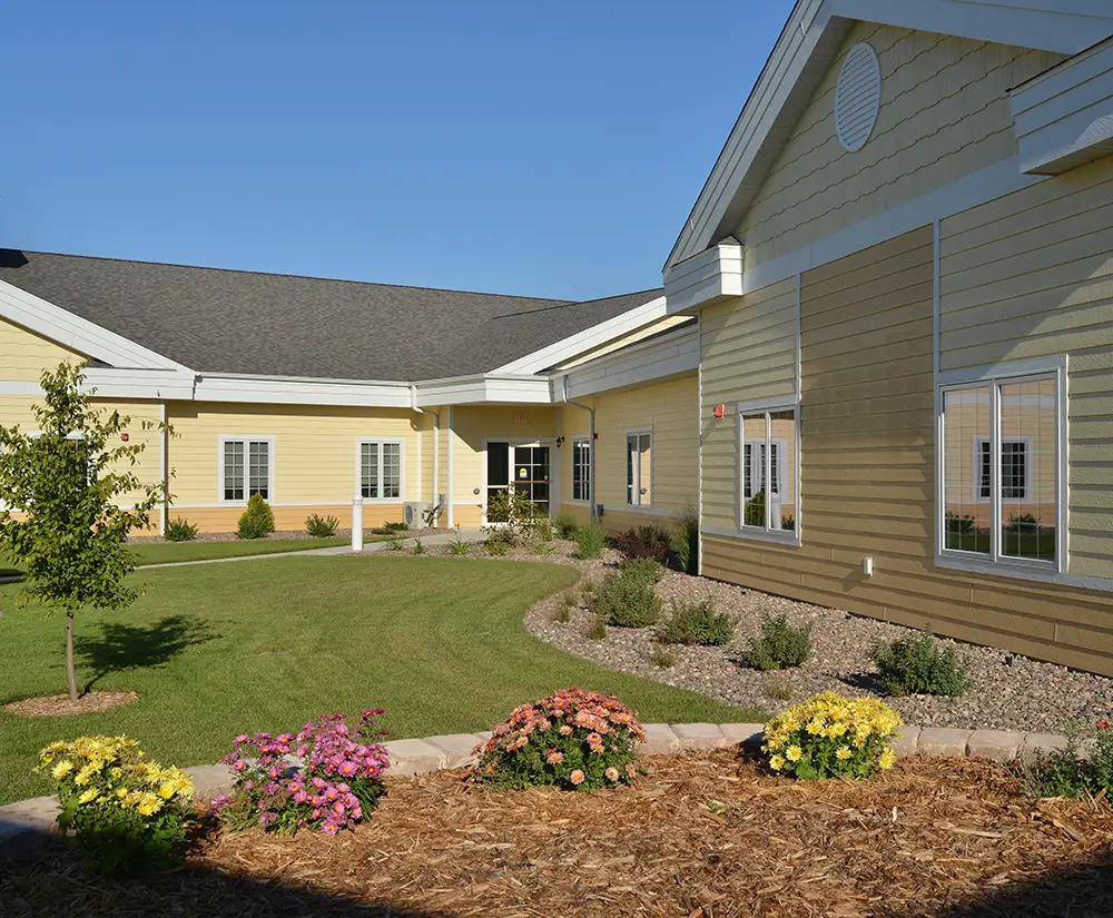 A single-story building with beige siding, surrounded by neatly landscaped gardens and colorful flowers, under a clear blue sky.