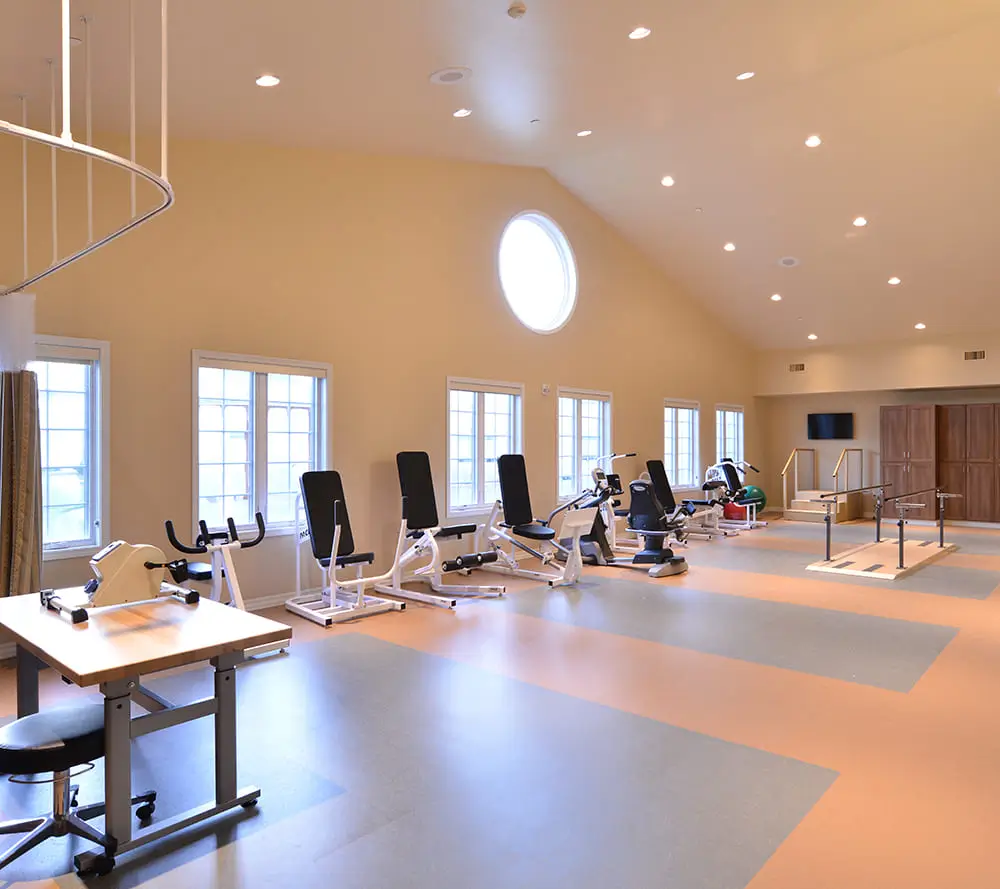 Spacious gym with exercise equipment, rehabilitation bars, and bright windows. Circular window on ceiling. No people present. Warm lighting, modern design.