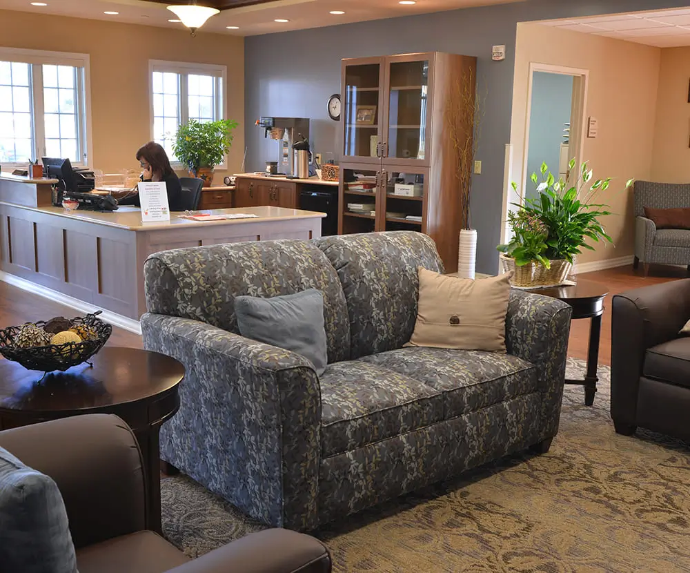 A cozy lobby features patterned sofas, a person at a reception desk, large windows, plants, and a coffee station in the background.