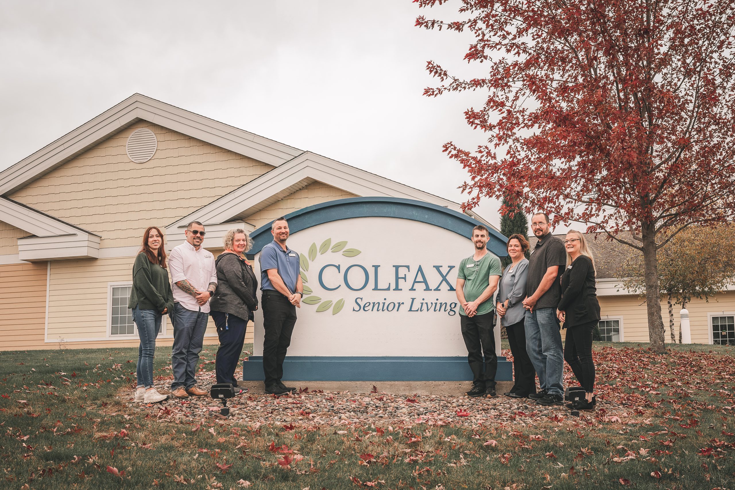 A group of people poses in front of a Colfax Senior Living sign outside a beige building with autumn leaves on the ground.