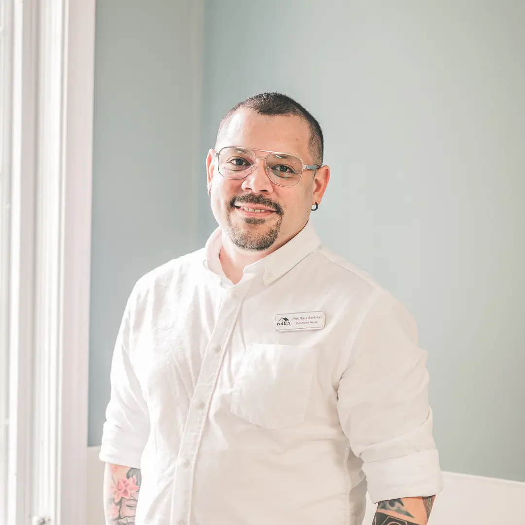 Smiling man with glasses and visible tattoos on his forearms, wearing a white button-down shirt with a name badge, standing indoors near a window.