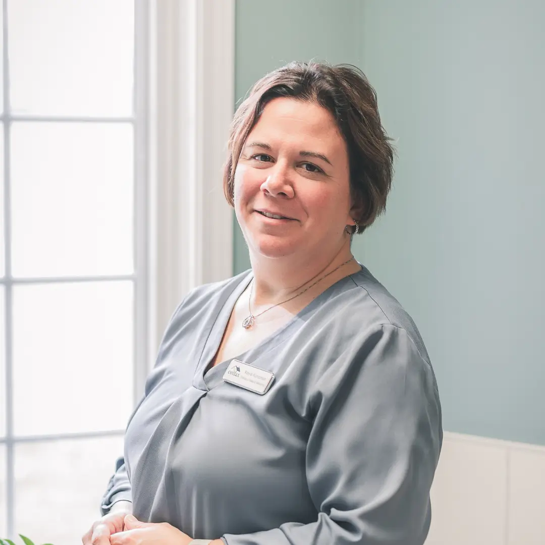 A smiling woman in gray scrubs with a name badge stands in a bright room by a window.