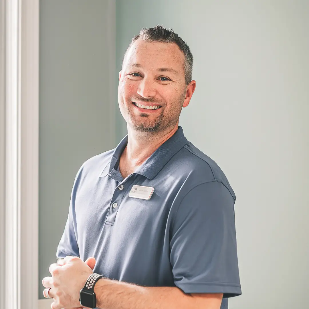 Smiling man in a blue polo shirt with a name badge stands indoors by a window.