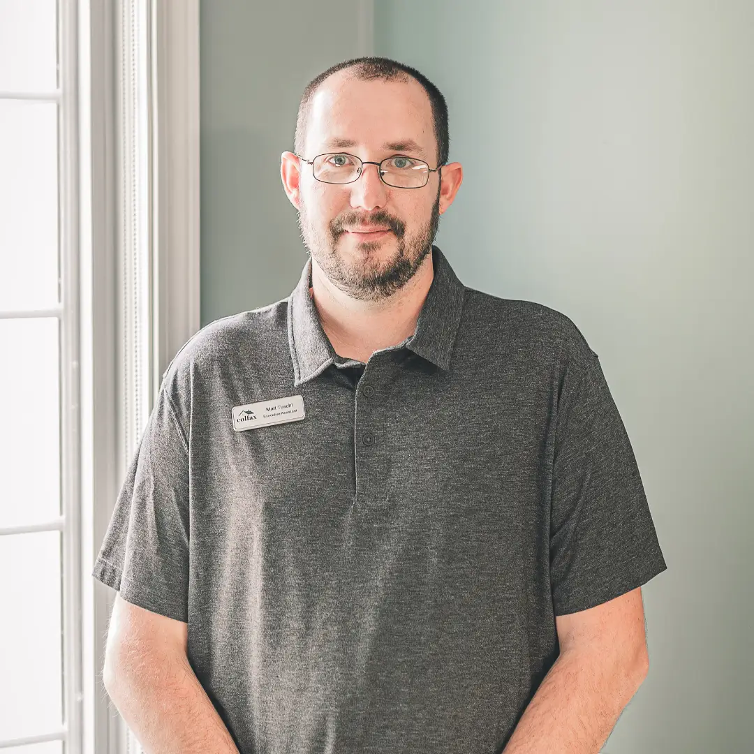 Man with glasses and a short beard wearing a gray polo shirt stands indoors by a window.