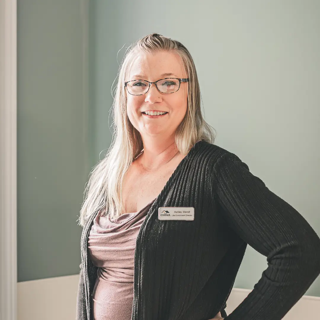 Smiling woman with glasses and long light hair wearing a name badge and a black cardigan over a mauve top, standing with one hand on her hip.
