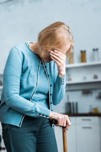 An elderly woman in a teal cardigan covers her face with one hand while leaning on a cane in a kitchen.