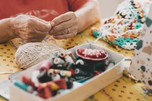 Close-up of a person&rsquo;s hands threading a needle beside a ball of yarn and a tray of colorful craft items on a yellow polka-dotted table.