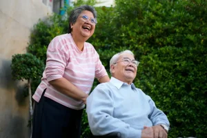 Smiling elderly woman pushes an elderly man in a wheelchair outdoors in a garden.