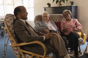 Three elderly people sit in upholstered armchairs in a cozy living room with a fireplace, chatting.