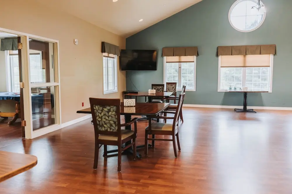 Bright dining area with hardwood floors, a wooden table and chairs, glass doors on the left, and windows with blinds along a teal-green wall.