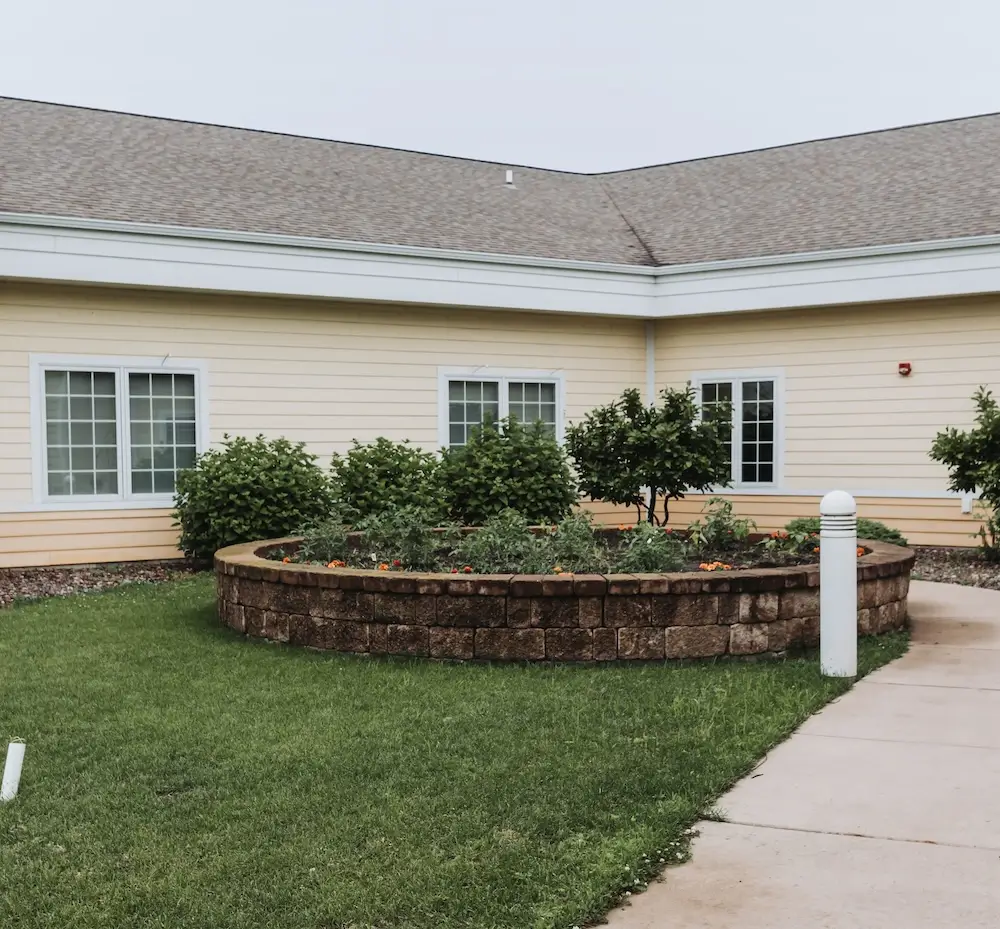 A single-story beige building with a circular brick planter filled with shrubs in front and a concrete sidewalk to the side.